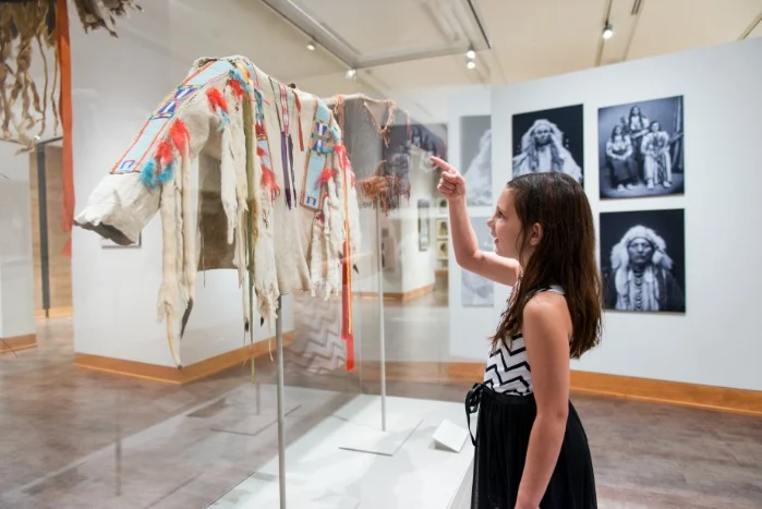 Meisje wijst naar een versierde leren mantel in een vitrine, met historische zwart-witfoto's van indianen aan de wand erachter