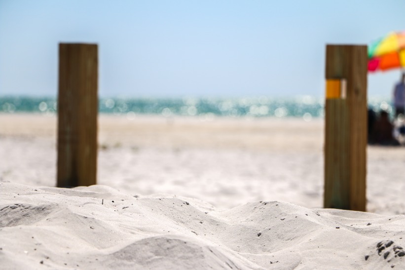 Twee houten paaltjes steken uit het fijne witte zand terwijl op de achtergrond zee en een strandparasol te zien zijn