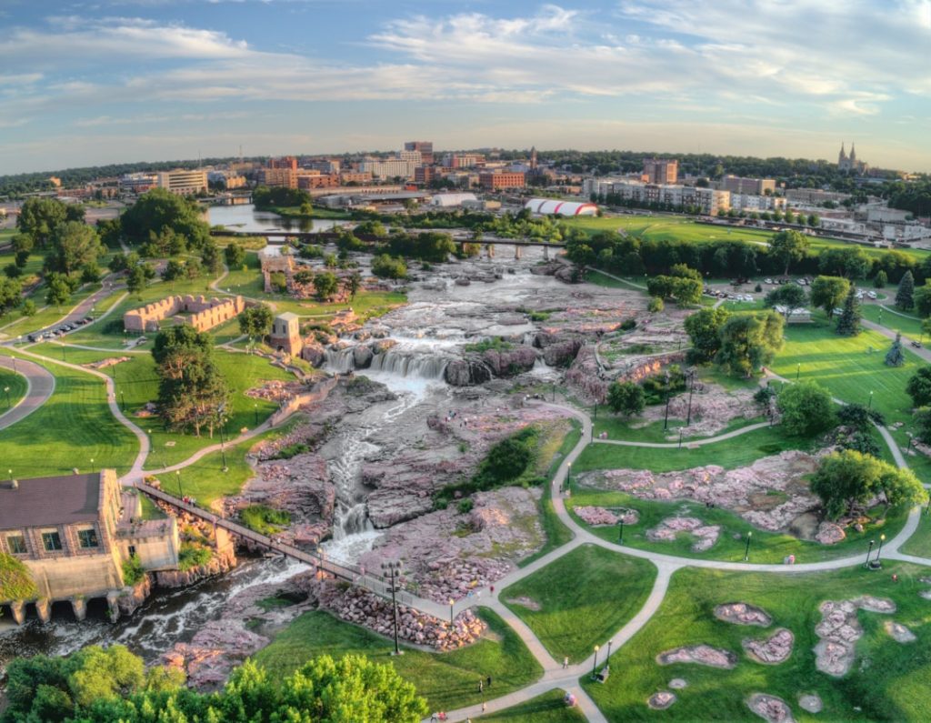 Waterval stroomt over roze kwartsietrotsen in een stadspark met wandelpaden en de skyline van Sioux Falls op de achtergrond