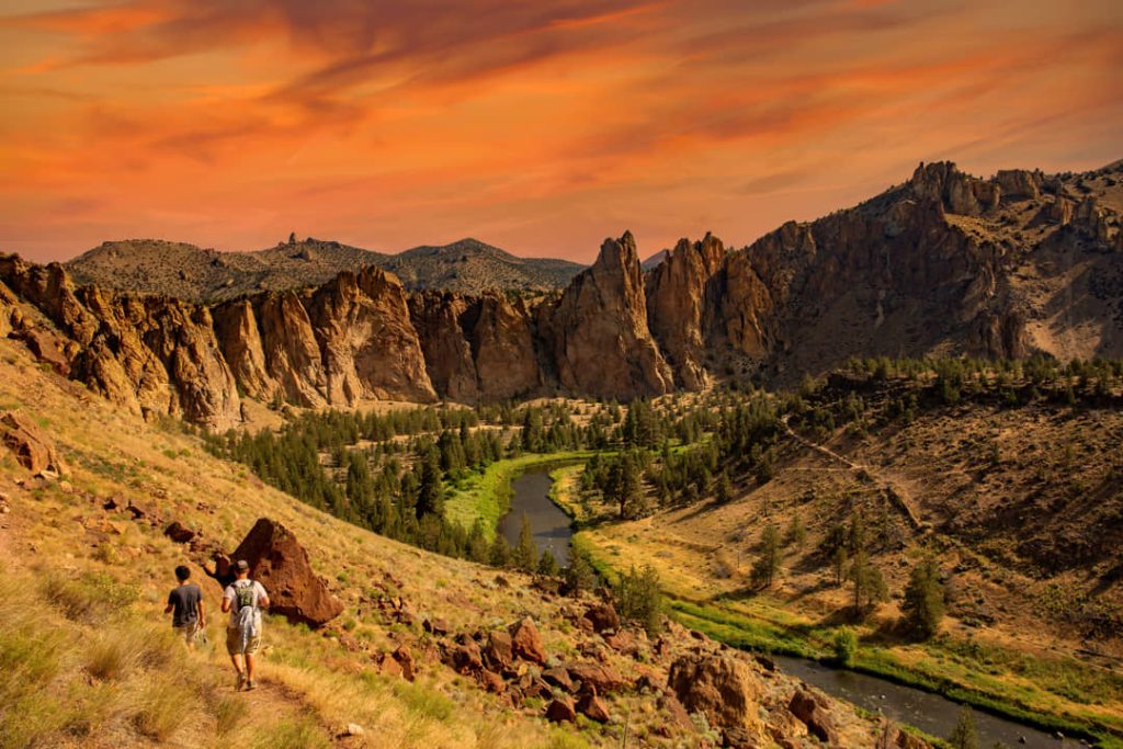 Twee wandelaars dalen een pad af tussen rotsformaties in Smith Rock State Park onder een dramatische rode avondlucht