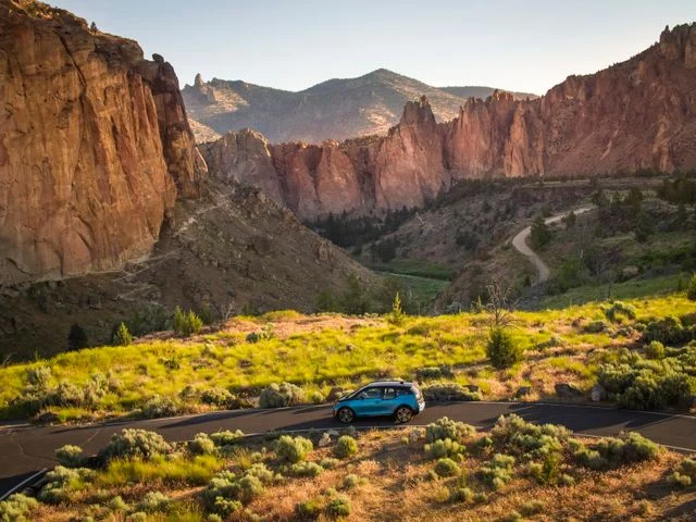 Kleine blauwe auto rijdt over een kronkelende weg door een vallei tussen hoge rode rotswanden in Smith Rock State Park in Oregon