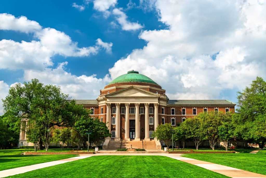 Bakstenen universiteitsgebouw met een groene koepel en zuilenportiek, gezien over een groot grasveld op de campus in Dallas