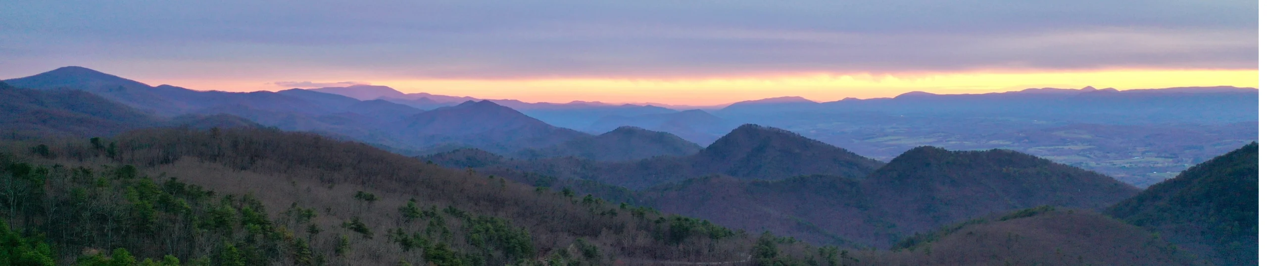 Panoramisch uitzicht over glooiende beboste bergketens in South Carolina met een zachte paars-oranje zonsondergang aan de horizon
