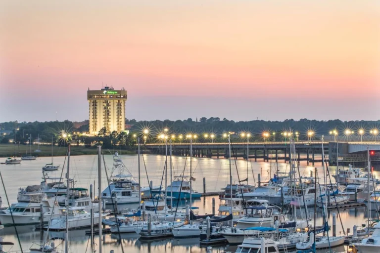 Een jachthaven vol boten bij zonsondergang in South Carolina, met een verlichte brug en het Holiday Inn hotel op de achtergrond