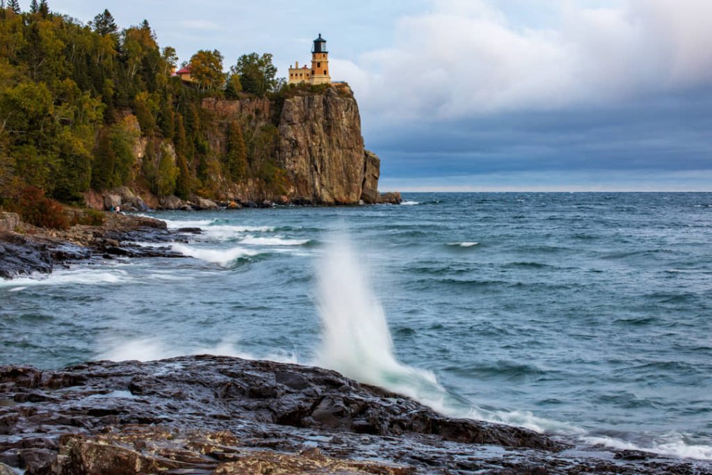 Vuurtoren Split Rock Lighthouse op een klif, met op de voorgrond een golf die hoog opspat tegen de rotsen