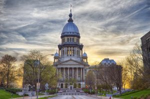Koepelgebouw van het Illinois State Capitol met verlichte zuilen en een vlag op de top, gefotografeerd vanaf de brede oprijlaan bij zonsondergang
