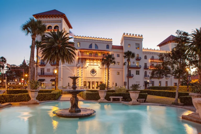 Het historische hotel Casa Monica in Saint Augustine met verlichte gevel, palmbomen en een fontein op de voorgrond bij schemering