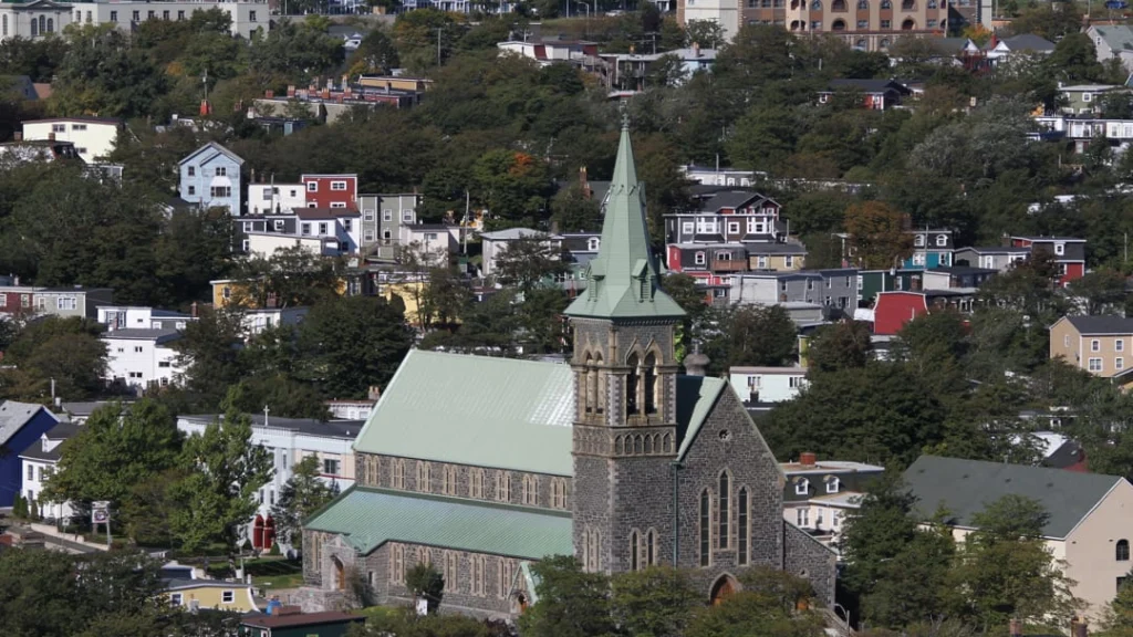 Stenen kerk met een hoge groene torenspits temidden van kleurrijke huizen op een heuvel in St. John's