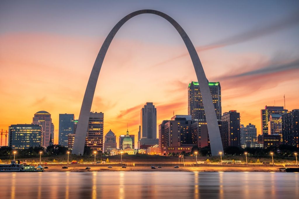 De grote stalen boog van de Gateway Arch tegen een oranje avondlucht, met de skyline van St. Louis en de rivier op de voorgrond
