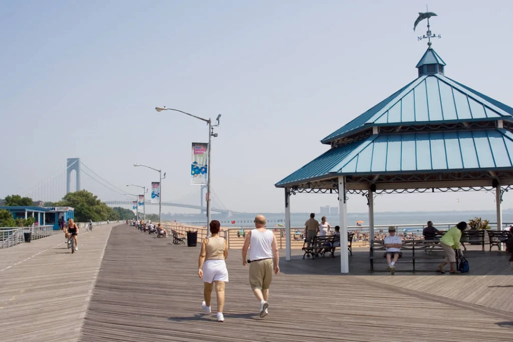 Wandelaars op een houten boardwalk langs het water met een blauw paviljoen en de Verrazzano Narrows Bridge op de achtergrond