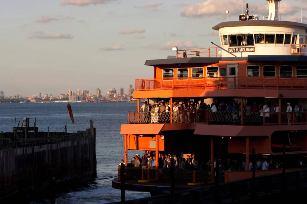 Passagiers aan boord van de oranje Staten Island Ferry bij zonsondergang met de skyline van Manhattan in de verte