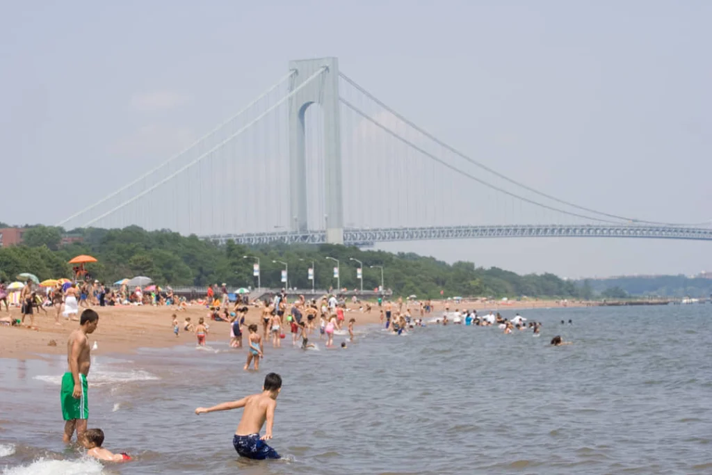 Mensen zwemmen en spelen op het zandstrand van South Beach terwijl de Verrazzano Narrows Bridge op de achtergrond oprijst