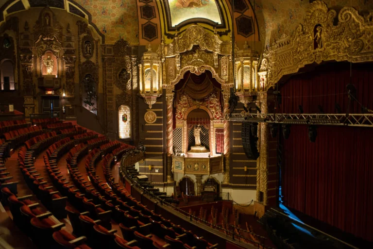Weelderig gedecoreerd balkon met rode stoelen en een gouden proscenium boven het podium in het St. George Theatre
