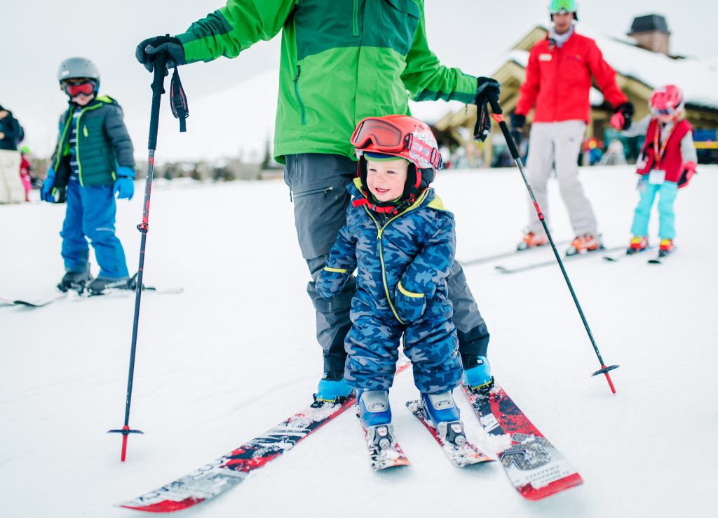 Kleine kinderen met helm en ski's staan op de sneeuw met begeleiders om hen heen tijdens een skiles op een beginnershelling