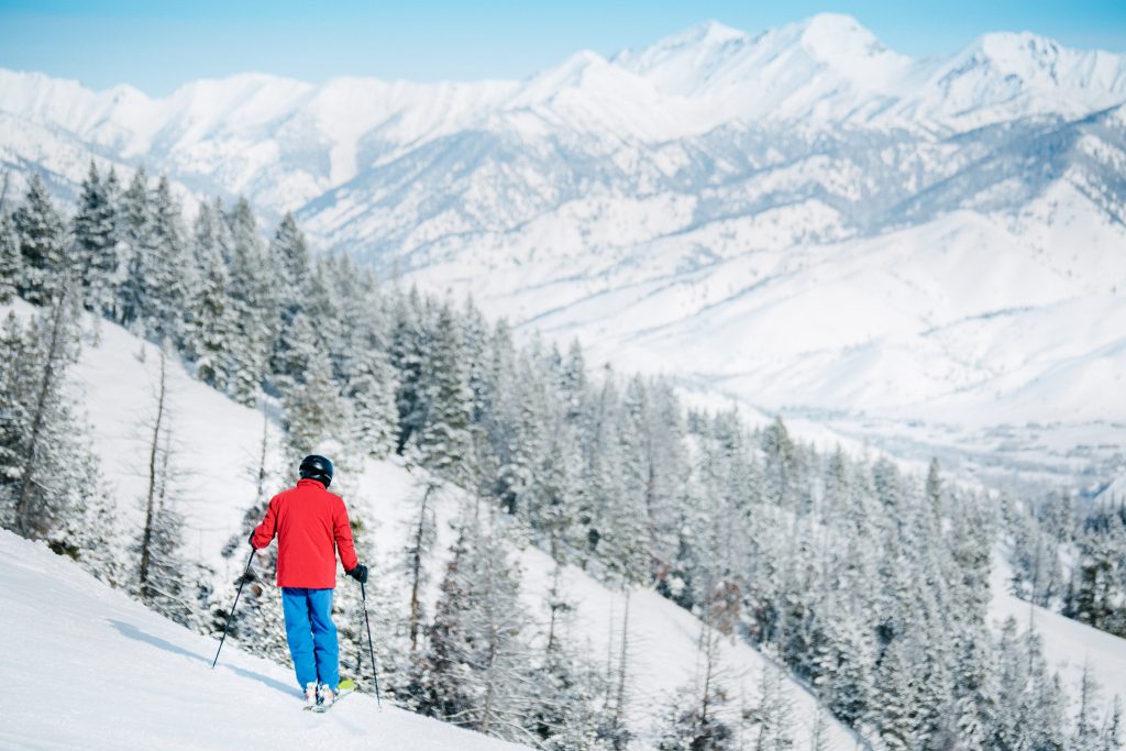 Skiër in rode jas en blauwe broek daalt af over een besneeuwde piste met dennenbos en een uitgestrekt bergpanorama op de achtergrond