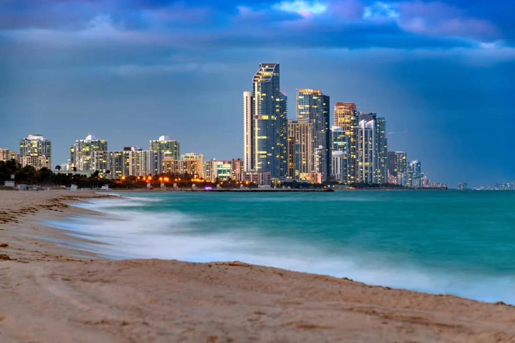 Verlichte hoogbouw van Sunny Isles Beach bij schemering gezien vanaf een breed zandstrand met golven op de voorgrond
