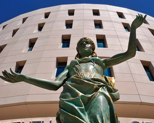 Bronzen vrouwenstandbeeld met geheven armen voor een rond gebouw met veel ramen tegen een blauwe lucht