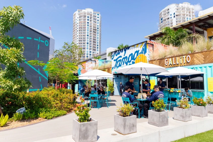 Bezoekers zitten onder parasols bij foodtrucks in een buitenpark met een grote muurschildering met de tekst Tampa