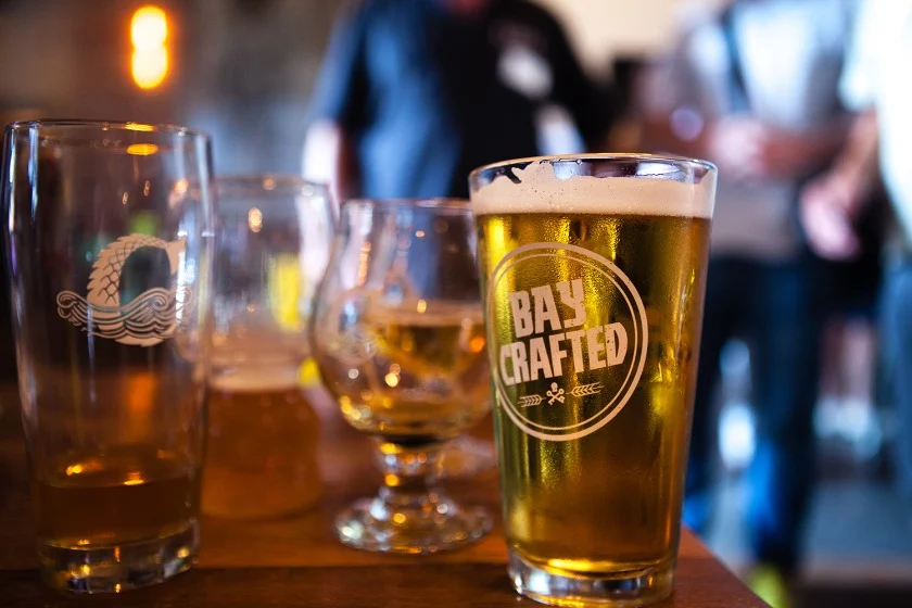 Close-up van een vol bierglas met het logo Bay Crafted naast andere proefglaasjes op een bar