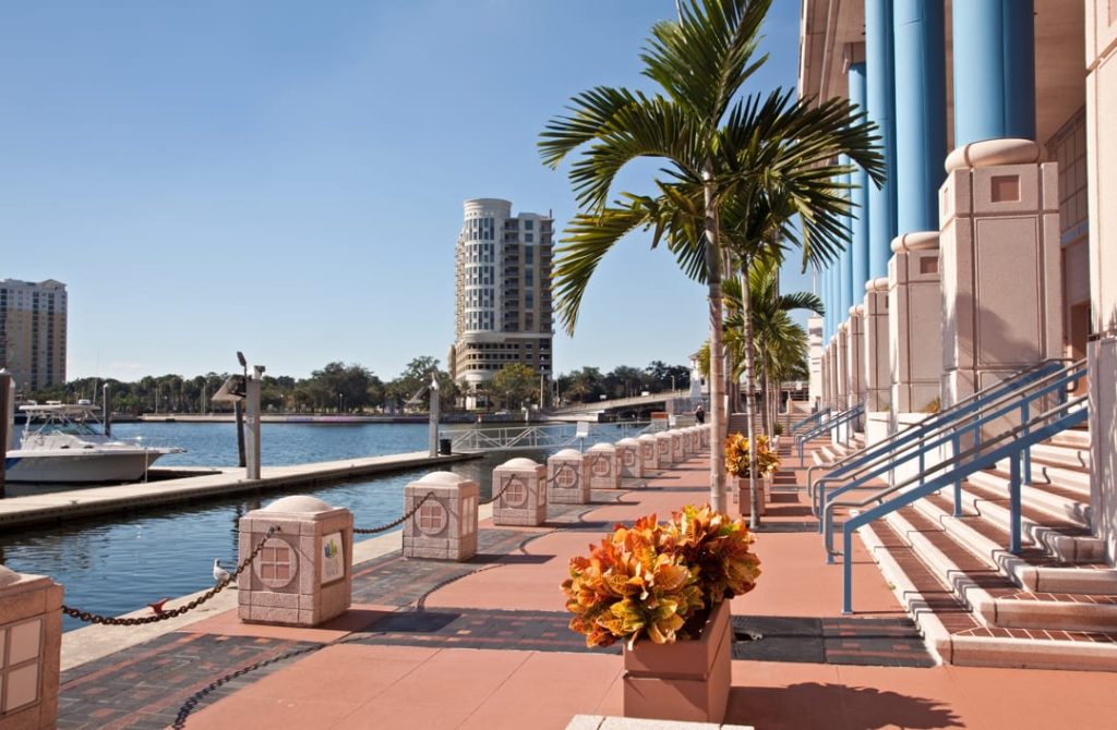 Bestrate rivierpromenade met een palmboom en bloemen naast een gebouw met blauwe zuilen, met de skyline op de achtergrond