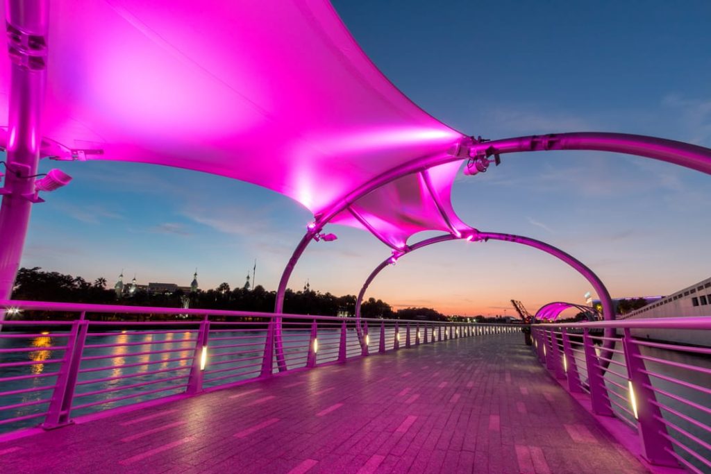 Overkapte voetgangersbrug met roze verlichting boven een pad langs het water in de avondschemering