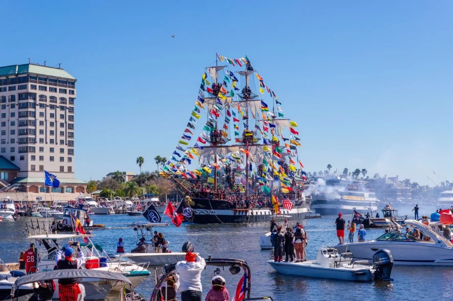 Een groot piratenschip versierd met kleurrijke vlaggen vaart door de haven van Tampa, omringd door toeschouwers in boten tijdens de Gasparilla-parade