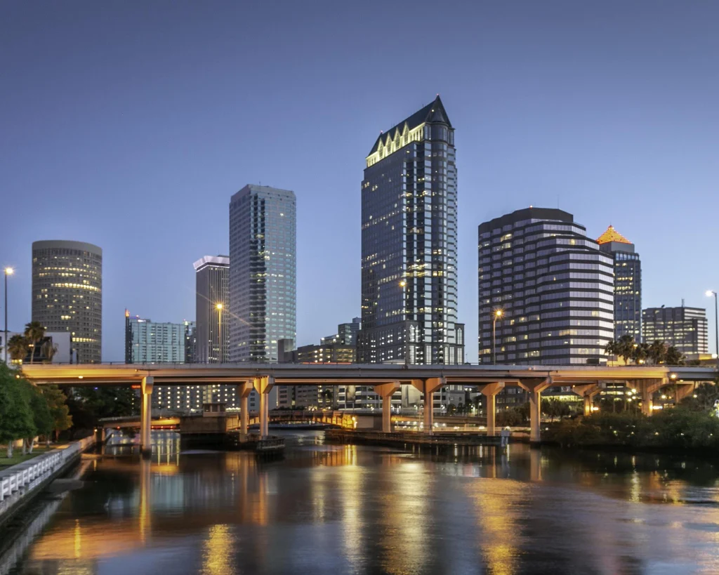 Verlichte wolkenkrabbers van de skyline van Tampa weerspiegeld in het water van de rivier, met een brug op de voorgrond bij schemering