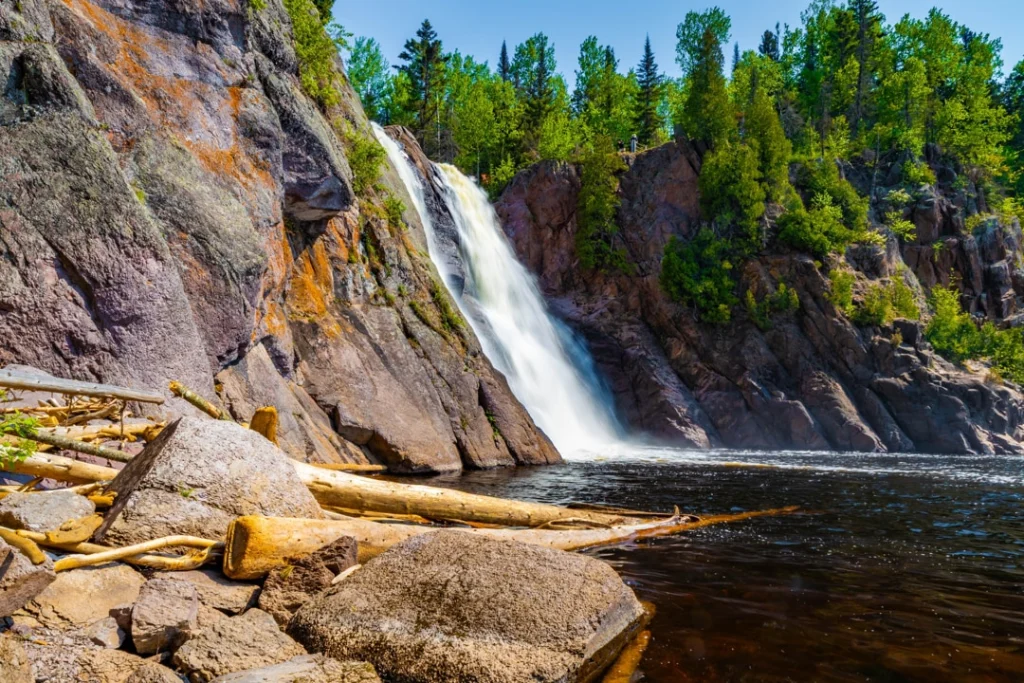 Brede waterval die over rotsen stort in Tettegouche State Park, met bos aan weerszijden en drijfhout op de voorgrond