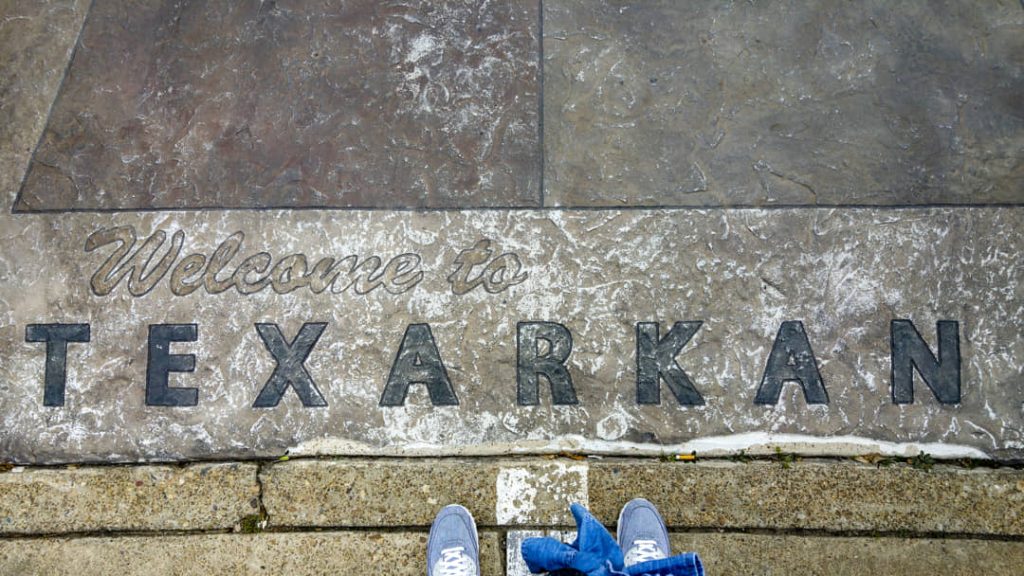 Tekst Welcome to Texarkana gegraveerd in een stenen stoep, met de voeten van een voorbijganger die op de grens staat