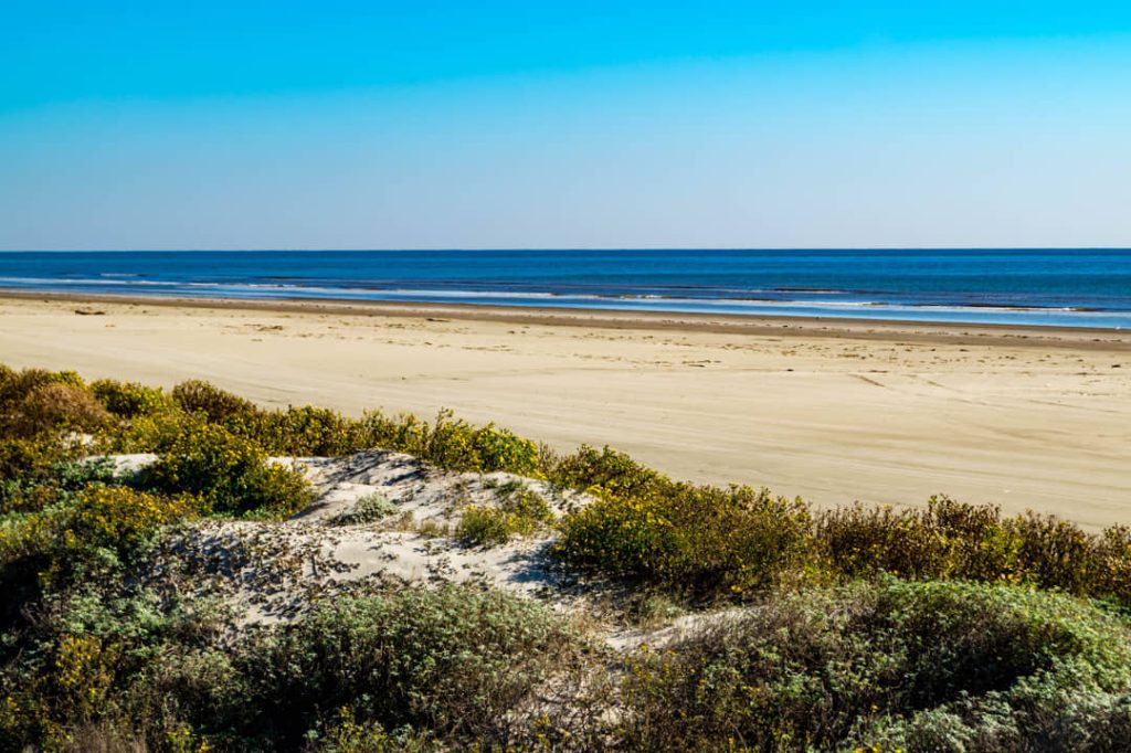 Breed leeg zandstrand met begroeide duinen op de voorgrond en kalme zee aan de golfkust van Texas onder een strakblauwe lucht