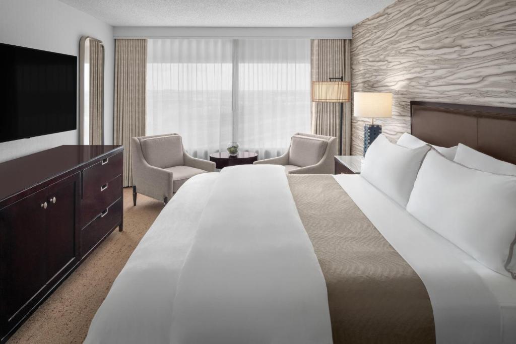 Hotelkamer met een groot bed en twee fauteuils bij een rond tafeltje voor het raam, met een dressoir en televisie
