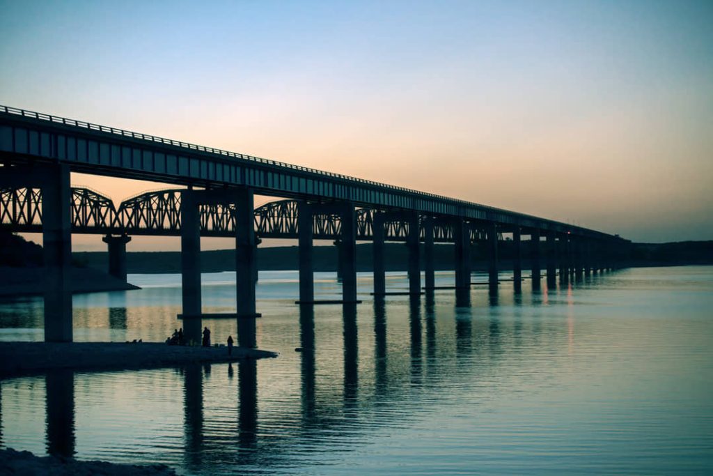 Silhouet van een lange stalen spoorbrug op pijlers over kalm water, weerspiegeld in het meer bij zonsondergang