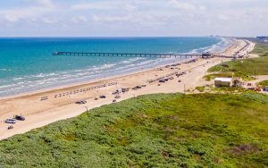 Luchtfoto van een zandstrand in Texas met een lange pier in zee, geparkeerde auto's en groene duinen op de voorgrond