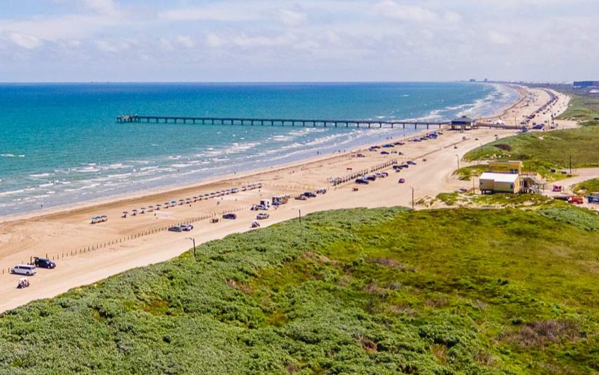 Luchtfoto van een zandstrand in Texas met een lange pier in zee, geparkeerde auto's en groene duinen op de voorgrond