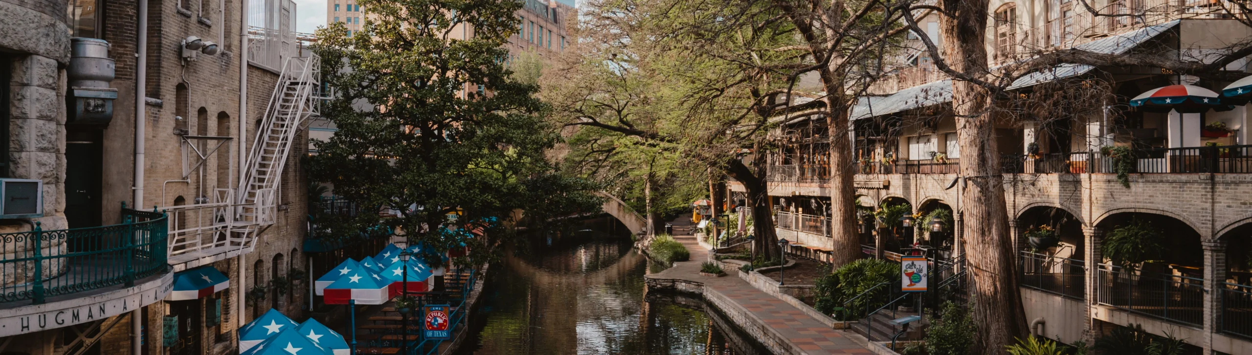 Uitzicht over de San Antonio Riverwalk in Texas met een rustig kanaal, blauw-witte parasols en historische gebouwen omringd door bomen