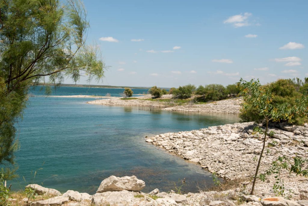Helder turquoise water van een meer met een rotsachtige, kale oever en struiken op de voorgrond onder een blauwe lucht
