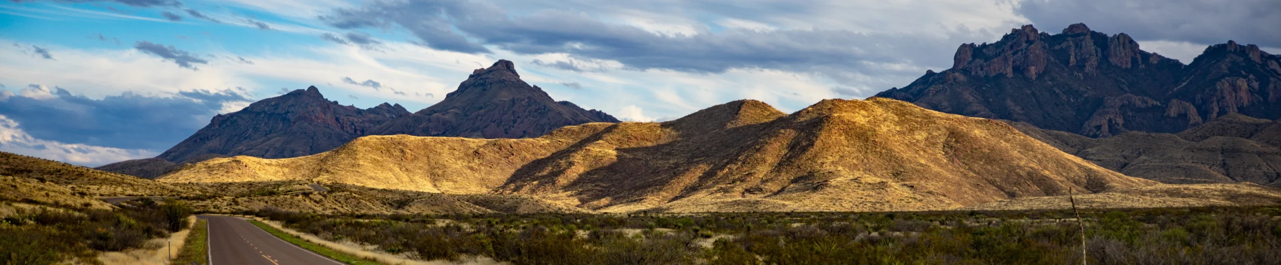Een lege asfaltweg slingert door een dorre woestijnvlakte in Texas met op de achtergrond een indrukwekkende bergketen onder een bewolkte lucht