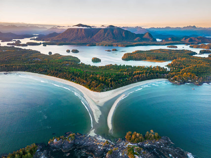 Luchtfoto van een smal zandstrand tussen twee baaien bij Tofino, omringd door beboste eilanden en bergen bij zonsondergang