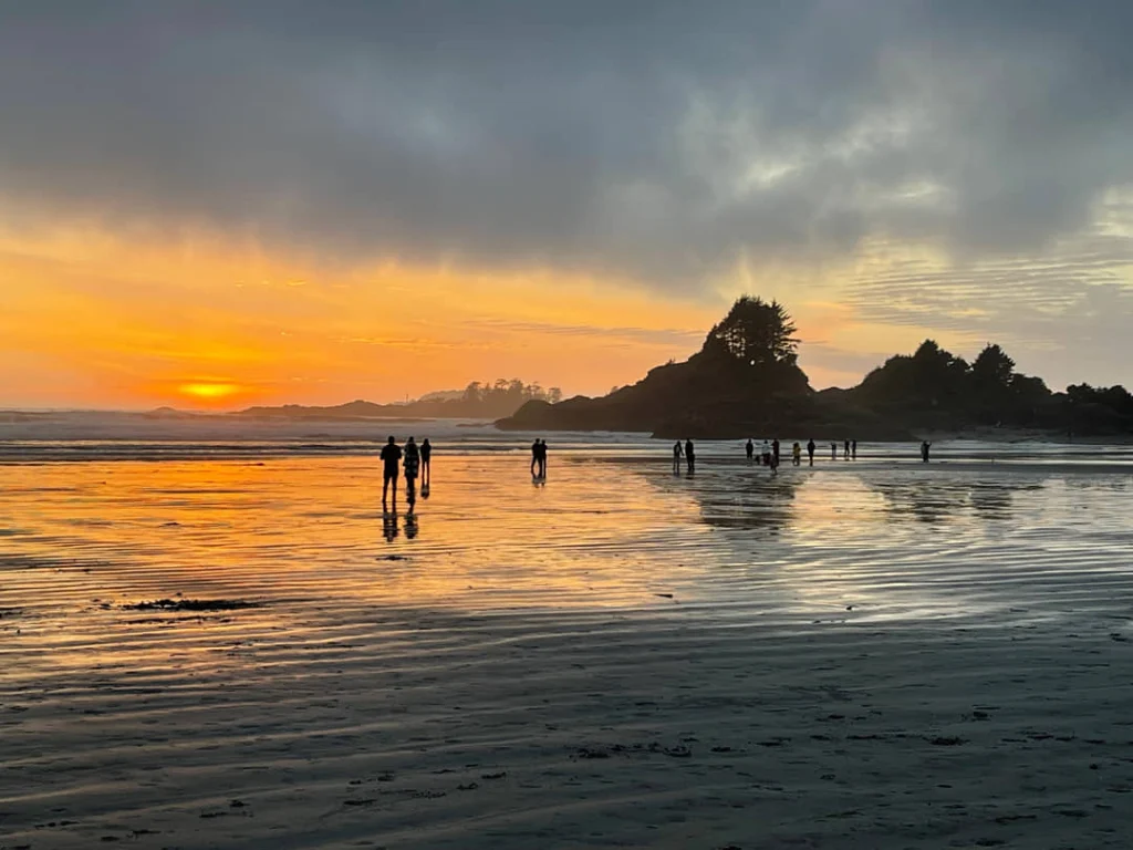 Silhouetten van mensen lopen over het natte strand van Tofino terwijl de zon oranje kleuren werpt over de horizon