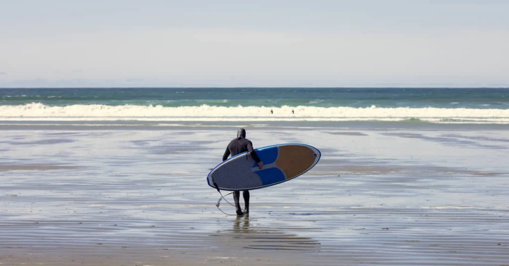 Een surfer in een wetsuit draagt een surfplank onder de arm terwijl hij over het natte strand van Tofino naar de golven loopt