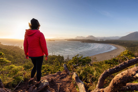 Vrouw in rode jas staat op een heuvel en kijkt uit over de kustlijn en baai bij Tofino tijdens zonsondergang