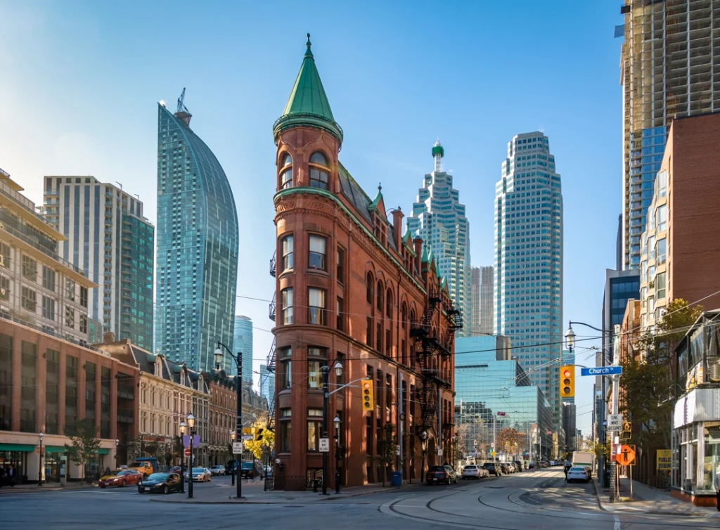 Rood bakstenen puntgebouw met groene torenspits op een drukke straathoek in het centrum van Toronto tussen moderne wolkenkrabbers