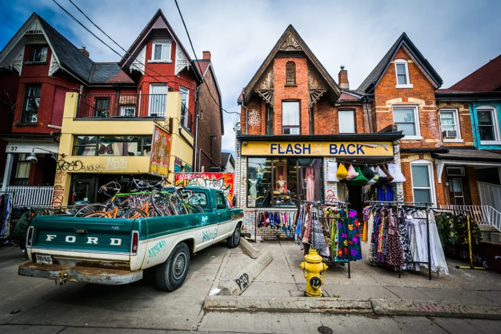 Straatbeeld in Kensington Market Toronto met een vintage kledingwinkel, een oude pick-up truck en kleurrijke kledingrekken buiten