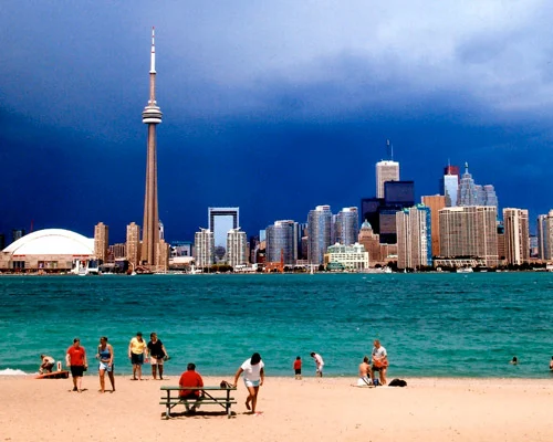 Mensen op een zandstrand aan het water met de skyline van Toronto en de CN Tower op de achtergrond onder een donkere onweerslucht