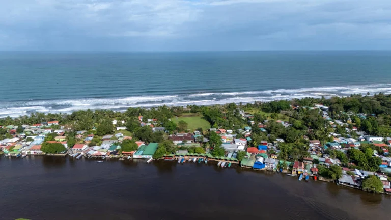 Luchtfoto van kleurrijke huisjes in het dorp Tortuguero met aan de ene kant een rivier en aan de andere kant golven van de oceaan