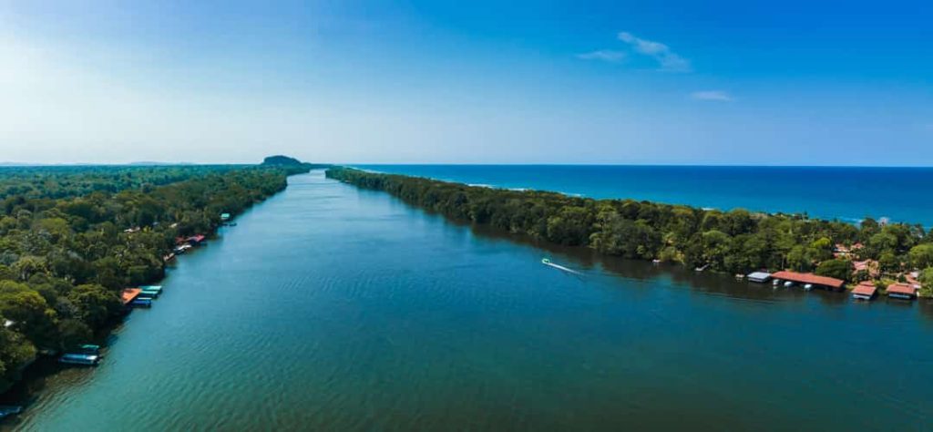 Luchtfoto van een breed blauwgroen kanaal tussen regenwoud met verspreide huizen langs beide oevers en de zee op de achtergrond