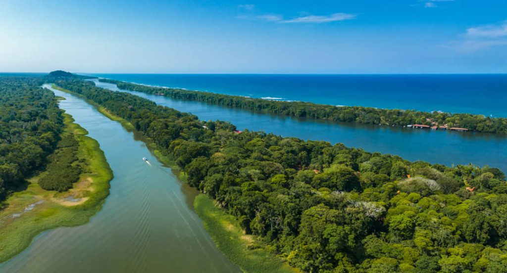 Luchtfoto van een breed waterkanaal dat door dicht regenwoud loopt, met de Caribische Zee zichtbaar aan de horizon