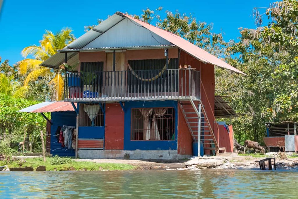 Tweeverdiepingen tellend rood en blauw houten huis met balkon en buitentrap, direct aan de oever van een kanaal met palmbomen