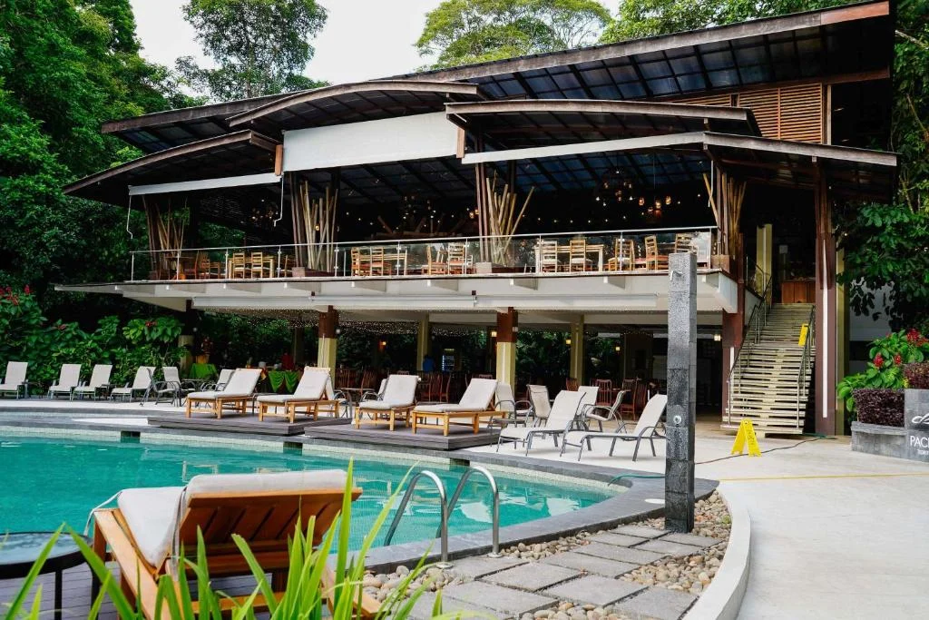 Een zwembad met ligstoelen ligt voor een open houten restaurant met rieten dak in het regenwoud bij een eco lodge in Tortuguero