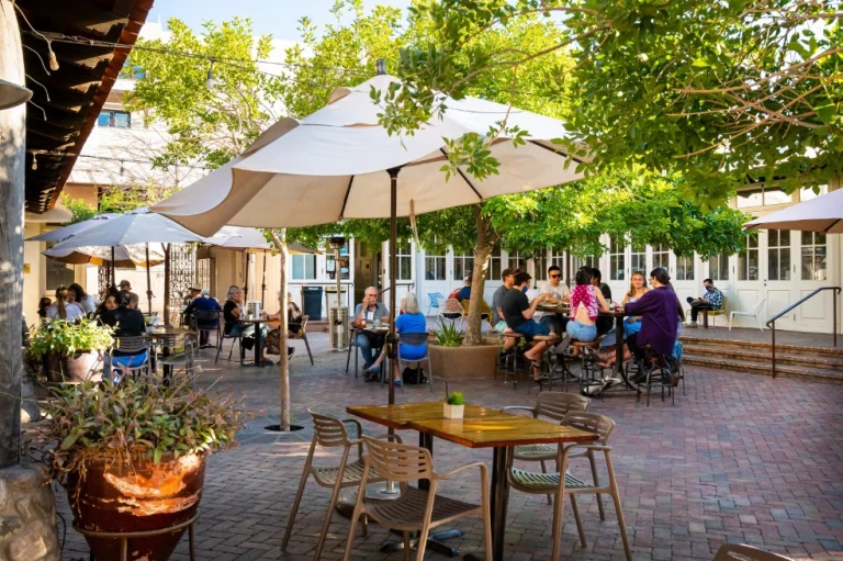 Mensen zitten aan houten tafels onder witte parasols op een schaduwrijk terras omringd door bomen in Tucson